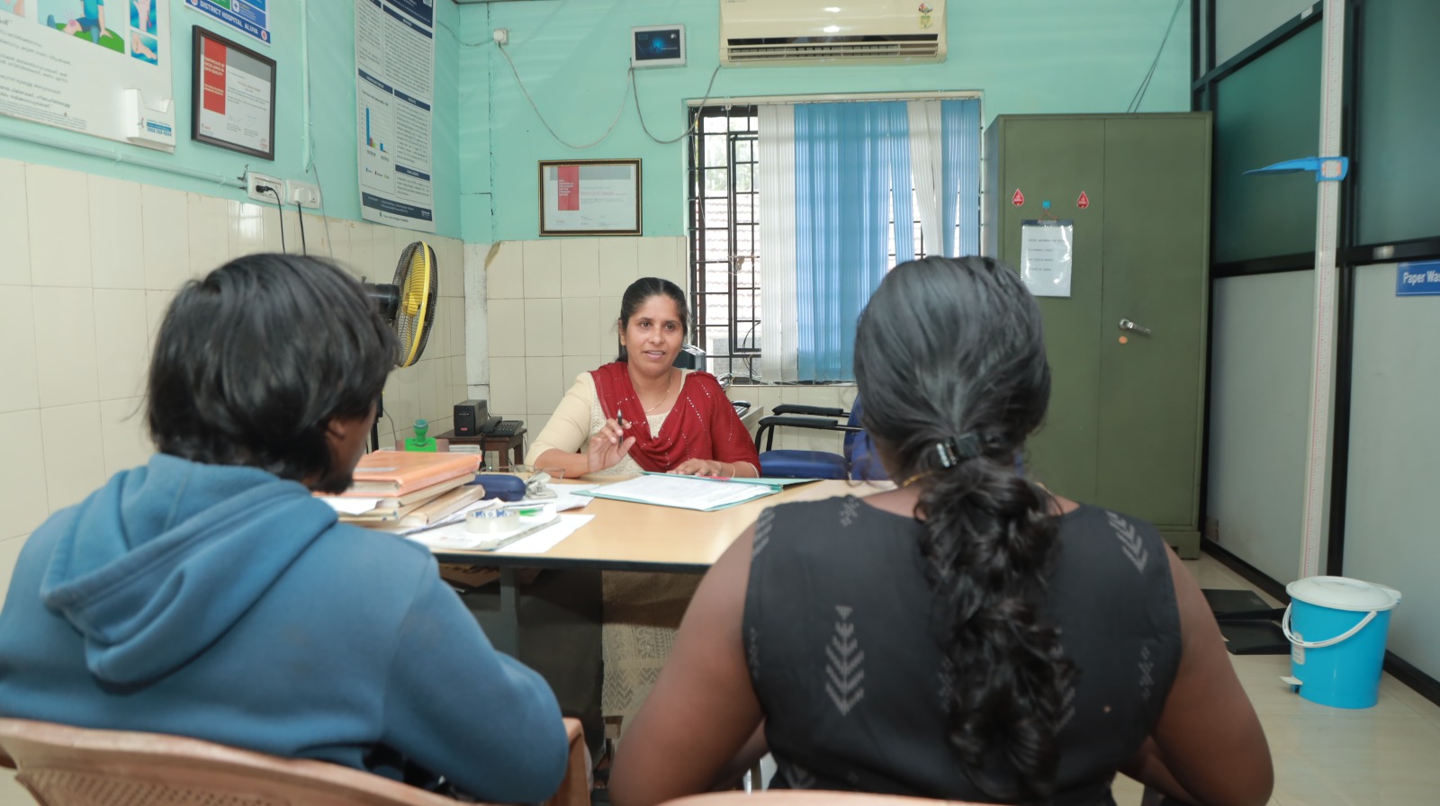 Indian doctors consulting Kerala family about hemophilia treatment with factor concentrate charts, traditional Kerala architecture with palm trees in background at District Hospital Aluva